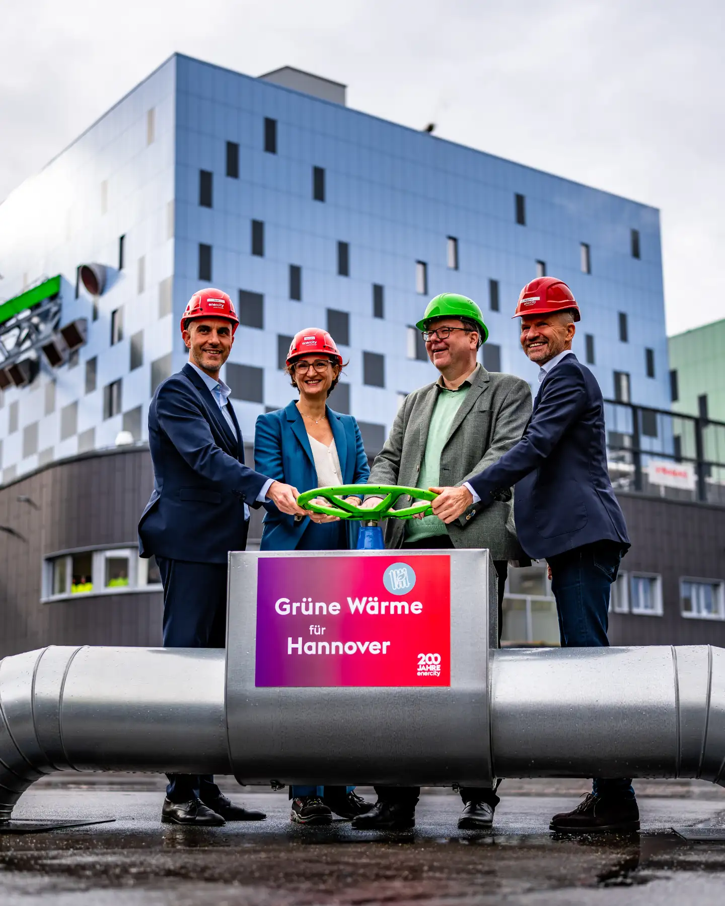 Vier Personen in Anzügen und Schutzhelmen stehen vor einem Industriegebäude und halten gemeinsam ein grünes Ventil; auf einem Schild steht „Grüne Wärme für Hannover“.