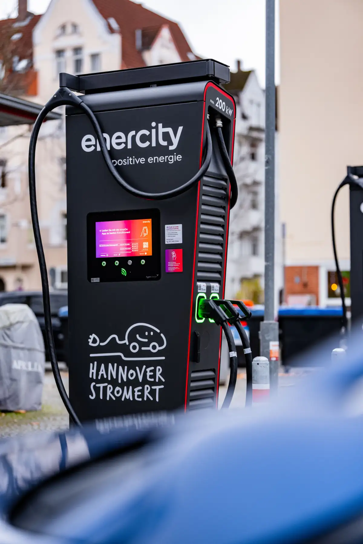 Ladepunkt für Elektroautos mit dem Schriftzug „enercity“ und „Hannover Stromert“, an dem mehrere Ladekabel angeschlossen sind, vor städtischer Häuserkulisse.